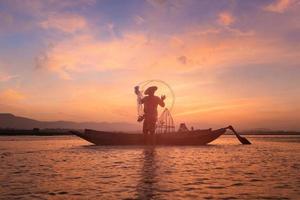 asiatisk fiskare på träbåt som kastar ett nät för att fånga sötvattensfisk i naturfloden tidigt på morgonen före soluppgången foto