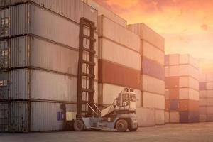 gaffeltruck lyfter lastcontainer i skeppsvarv eller hamngård mot soluppgångshimlen med lastcontainerstapel i bakgrunden för transport import, export och logistisk industrikoncept foto
