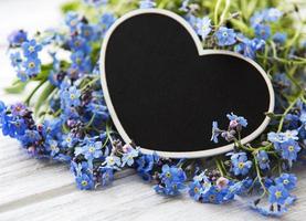 glöm mig-inte-blommor och svart hjärtformat bräde på vit träbakgrund foto