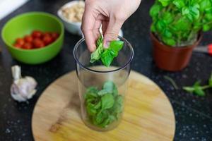 personen som kastar färska basilikablad i en mixer. beredning av en italiensk maträtt. basilika pesto. foto