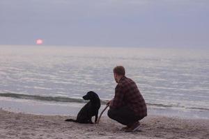 ung man går på morgonstranden med svart hund foto