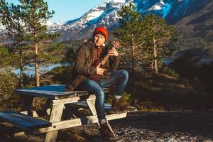 ung man spelar gitarr sitter på ett träbord mot bakgrund av berg, skogar och sjöar. koppla av och njuta av soliga dagar. plats för text eller reklam foto