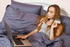 vacker kvinna shopping online med kreditkort och bärbar dator medan liggande på sängen hemma isolerade foto