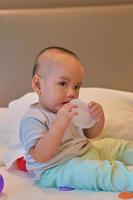 porträtt av glad 6 månader gammal asiatisk pojke sitter på sängen och spelar foto
