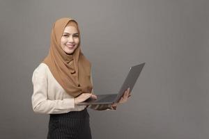 porträtt av vacker kvinna med hijab håller dator bärbar dator på grå bakgrund foto