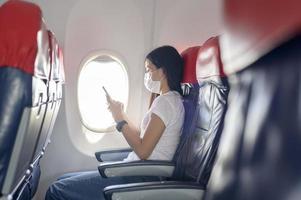 resande kvinna som bär skyddsmask ombord i flygplanet med smartphone, resor under covid-19-pandemin, säkerhetsresor, socialt avståndsprotokoll, nytt normalt resekoncept