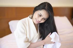 leende ung kvinna som bär vit morgonrock torkar håret med handduk efter en dusch i sovrummet foto