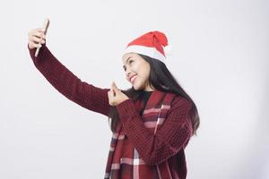 ung leende kvinna som bär röd jultomtehatt tar en selfie på vit bakgrund studio. foto