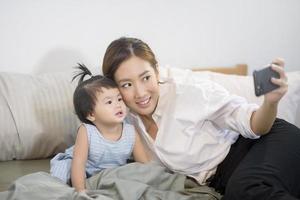 asiatisk mamma och hennes babydotter ringer selfie eller videosamtal till pappa i sängen, familj, hemsäkerhet, föräldraskap, teknikkoncept foto