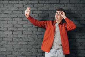 ung glad man i röd skjorta och glasögon tar selfiefoto på mobiltelefon på en bakgrund av svart tegelvägg. kopiera, tomt utrymme för text foto