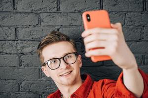 porträtt ung man i röd skjorta och glasögon tar selfiefoto på mobiltelefon på en bakgrund av svart tegelvägg. kopiera, tomt utrymme för text foto