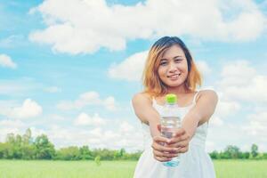 kvinnors livsstilskoncept ung vacker kvinna som bär vit klänning som håller en flaska vatten på gräsmarkssmiley till kameran ser så fräsch ut, njut av och glad. foto