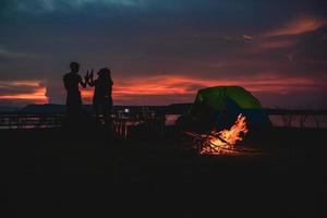 siluett grupp av asiatiska vänner turist dricker och spelar gitarr tillsammans med lycka på sommaren medan du har camping nära sjön foto