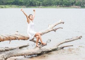 ung kvinna sitter sträcka händerna på trädstammen vid sjön och ler. foto