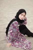 vacker islamisk kvinnlig modell som bär hijabmode, en modern bröllopsklänning för muslimsk kvinna som sitter i sanden och stranden. porträtt av en asiatisk tjejmodell med hijab som har kul på stranden med träd foto