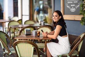 leende kvinna med blå ögon sitter på urban café med smart telefon foto
