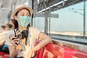 vacker asiatisk kvinnlig turist med ansiktsmask sitter i ett rött säte, reser med tåg, tar ögonblicksbilder, transporterar i förortsvy, njuter av passagerarlivsstil med järnväg, lycklig semester. foto