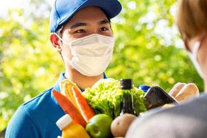 hygienisk asiatisk budbärare som bär ansiktsmask medan han levererar matvaror till kunden hemma, matleverans i pandemikonceptets tid foto