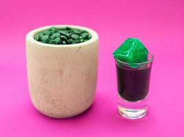 flytande spirulina grön drink i cocktailglas och spirulina piller på rosa bakgrund. supermat, hälsosam livsstil, hälsosamt kosttillskott koncept foto