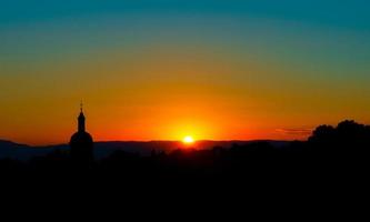 den fantastiska skönheten och färgerna i solnedgången med utsikt över alpernas silhuetter och silhuetten av en vacker kyrka foto