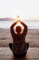 ung frisk kvinna som utövar yoga och håller händerna i meditationsställning med solen mellan dem på stranden vid soluppgången foto