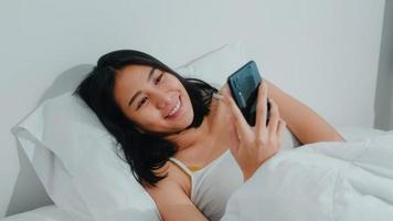 ung asiatisk kvinna som använder smartphone och kollar sociala medier känner sig glad leende när hon ligger på sängen efter att ha vaknat på morgonen, vacker attraktiv spansktalande dam som ler slappna av i sovrummet hemma. foto