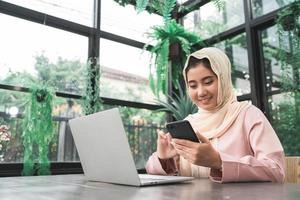 vacker ung leende asiatisk muslimsk kvinna som arbetar på telefonen sitter i vardagsrummet hemma. Asiatisk affärskvinna arbetsdokument finans och kalkylator i hennes hemmakontor. njuta av tiden hemma. foto