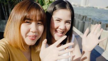 lesbisk hbtq kvinnor influencer par viftande hand ringer videosamtal på café. asiatisk älskare dam glad slappna av kul med teknik telefon spela in vlogg-videouppladdning i sociala medier resa nära floden i staden. foto