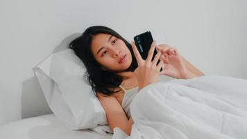 ung asiatisk kvinna som använder smartphone och kollar sociala medier känner sig glad leende när hon ligger på sängen efter att ha vaknat på morgonen, vacker attraktiv spansktalande dam som ler slappna av i sovrummet hemma. foto