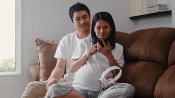 unga asiatiska gravida par som använder telefon och hörlurar spelar musik för baby i magen. mamma och pappa känner sig glada leende fridfulla medan ta hand om barn som ligger på soffan i vardagsrummet hemma koncept. foto