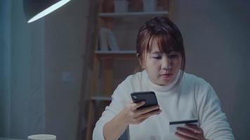 vacker asiatisk kvinna som använder smartphone och köper onlineshopping med kreditkort medan du bär avslappnad sittande på skrivbordet i vardagsrummet på natten hemma. livsstil kvinnor som arbetar hemma kontor koncept. foto