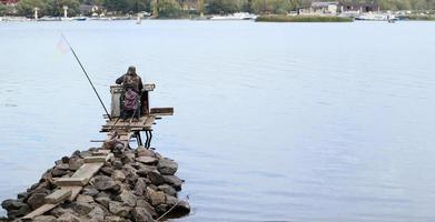 en fiskare fångar fisk på floden, bakifrån, från stranden. en fiskare sitter på en trä- och stenbro på flodstranden och försöker fånga fisk. sport, rekreation, livsstil foto