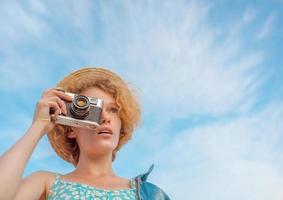 ung lockig rödhårig kvinna i stråhatt, blå sundress och jeansjacka som står med vintagekamera och tar bilder på blå himmel bakgrund. kul, sommar, mode, fotografering, resor, ungdomskoncept foto