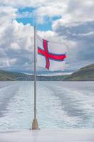 Färöarnas nationella flagga fladdrar i vinden fäst vid en turistbåt vid Färöarnas skärgård och Atlanten, sommar. foto