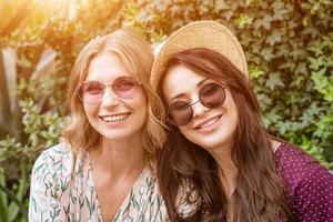 porträtt av två glada kvinnor i solglasögon och hattar på en bakgrund av lövverk foto