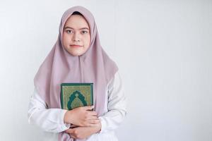 ung asiatisk islam kvinna som bär huvudduk håller den heliga Koranen med nära ögon och lugnt ansikte. indonesisk kvinna på grå bakgrund foto