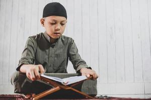 asiatisk muslimsk pojke som bär traditionell dräkt som kallas gamis och läser den heliga boken al-Koranen på bönemattan foto