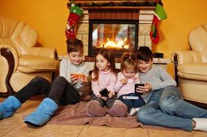 glada fyra barn tittar på i mobiltelefonen hemma vid en öppen spis i varma vardagsrummet på vinterdagen. foto