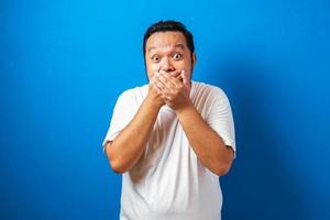 fet asiatisk man i vit t-shirt visar roliga chockade och överraskande uttryck mot blå bakgrund foto