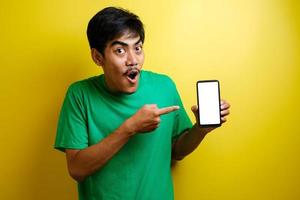 porträtt av chockad ung asiatisk man i grön t-shirt pekar på mobiltelefon foto