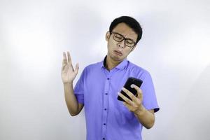 ung asiatisk man gråter och ledsen när han tittar och använder telefonen. indonesien man bär glasögon och blå skjorta isolerade blå bakgrund. foto