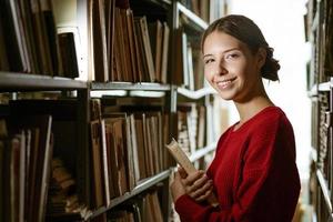 flickan håller en bok i sina händer mot bakgrunden av biblioteket. foto