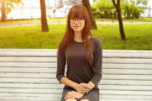 porträtt av en söt tjej med glasögon som sitter på en bänk på gatan i parken foto