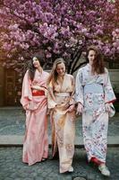 tre europeiska flickor som bär traditionell japansk kimonobakgrund blommar rosa sakuraträd foto