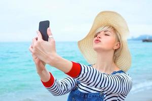 vacker ung blond kaukasisk kvinna på semester i stråhatt, randig blus nad jeansoveraller gör selfie på smartphone vid den fantastiska blå havsbakgrunden foto