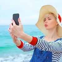 vacker ung blond kaukasisk kvinna på semester i stråhatt, randig blus nad jeansoveraller gör selfie på smartphone vid den fantastiska blå havsbakgrunden foto