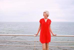 porträtt av ganska cool vackert kort hår blond kvinna i röd korallklänning på havsbakgrunden på stranden foto