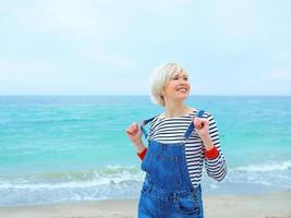 vacker ung blond kaukasisk kvinna på semester i randig blus, sneakers och jeansoverall på stranden vid den fantastiska blå havets bakgrund. glad att vara vid havet foto