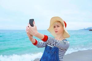 vacker ung blond kaukasisk kvinna på semester i stråhatt, randig blus nad jeansoveraller gör selfie på smartphone vid den fantastiska blå havsbakgrunden foto