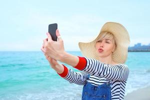 vacker ung blond kaukasisk kvinna på semester i stråhatt, randig blus nad jeansoveraller gör selfie på smartphone vid den fantastiska blå havsbakgrunden foto